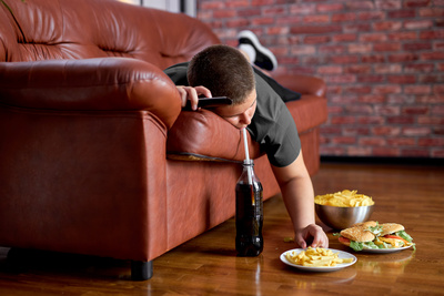 Übergewichtiger Junge liegt schlapp auf einem Sofa, trinkt COla und isst Fast Food