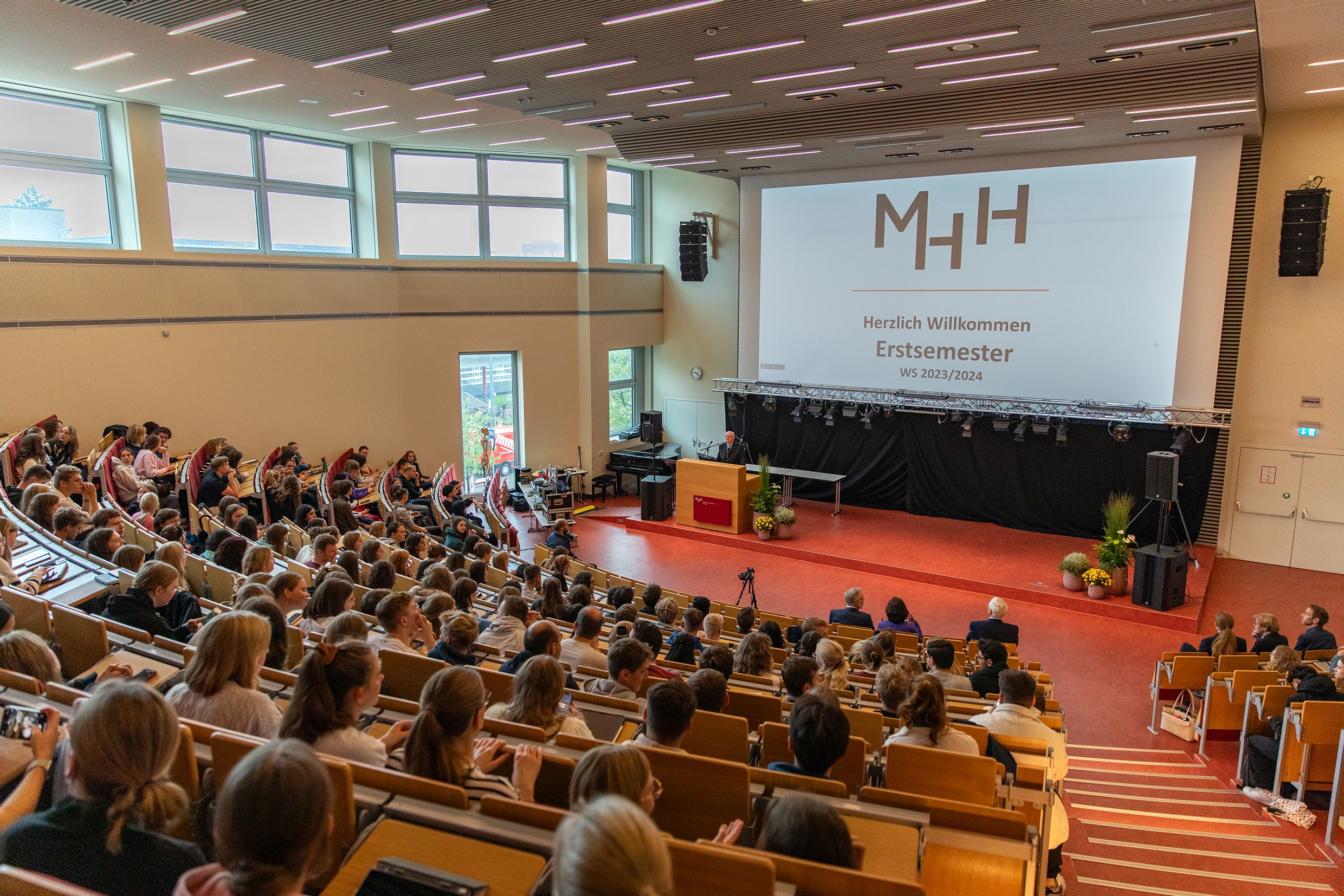 Das Foto zeigt die Erstsemesterbegrüßung der Medizinischen Hochschule Hannover. Es ist ein größer Hörsaal zu sehen, in dem die neuen Studierenden sitzen. Vorne an der Leinwand wird die Begrüßungspräsentation angezeigt. 